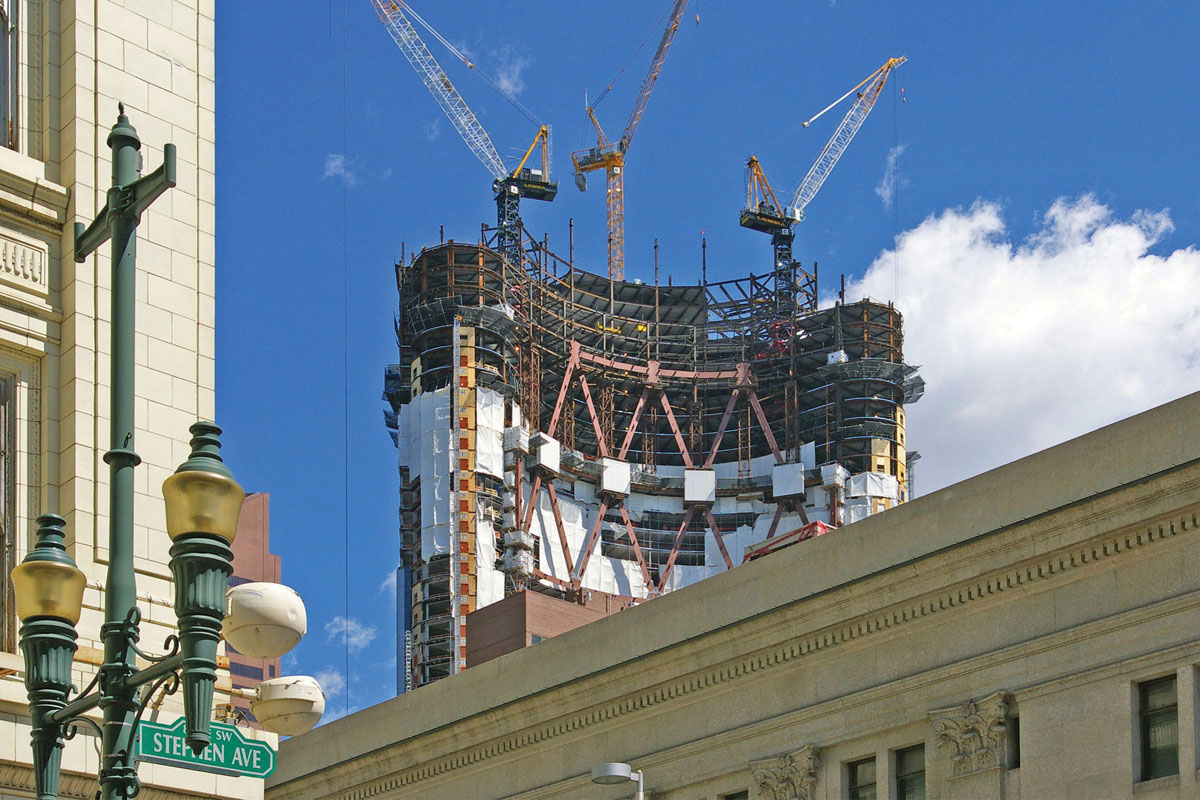 A phoograph of a curved skyscraper under construction, with traditional stone buildings and a vintage street light in the foreground.